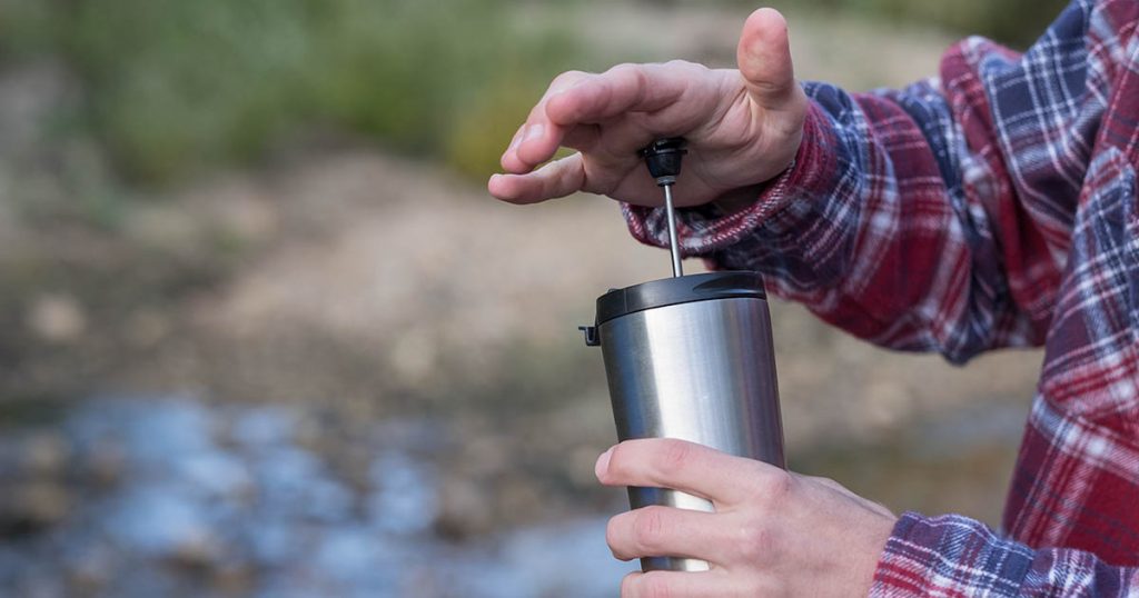 Pushing the plunger down on a French Press travel mug while camping by a river..
