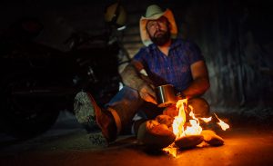 A man in a cowboy hat sitting next to a camp fire drinking cowboy coffee.